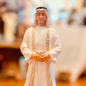 May include: A figurine of a man in traditional Arab dress. The figure wears a white robe with gold trim and a white headdress. The man's hands are clasped. The figurine has a detailed face and beard. The robe has decorative gold edging.
