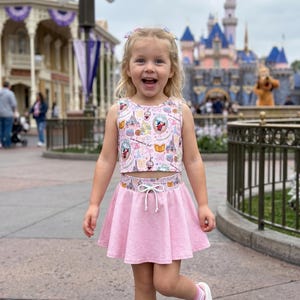 Può includere: Un completo rosa per bambina composto da crop top e gonna con coulisse bianca. Il top presenta un motivo colorato con immagini di cartoni animati. La bambina indossa scarpe da ginnastica bianche e calzini a righe rosa e bianche. Sullo sfondo c'è un castello.