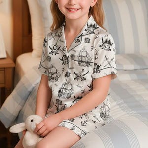 May include: A child wearing a white pirate-themed pajama set with black illustrations of ships and skulls. The sleepwear includes a short-sleeved top and shorts. The child is holding a soft toy bunny.