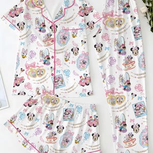 May include: A white pajama set with a pattern of Minnie Mouse, Daisy Duck, and other Disney characters. The set includes a short-sleeved button-up shirt, shorts, and long trousers, all trimmed with pink piping. The print includes cars, teacups, and bows.