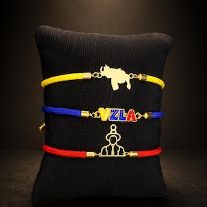 May include: Three cord bracelets with gold-tone metal charms. The top yellow cord features a map of Venezuela. The middle blue cord displays the letters VZLA in yellow, blue, and red. The bottom red cord features a gold-tone outline of a person.