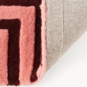 May include: Close-up of a pink and burgundy geometric patterned rug. The rug has a soft, textured surface with a light pink border and a neutral-colored backing. The design features a repeating pattern of burgundy lines.