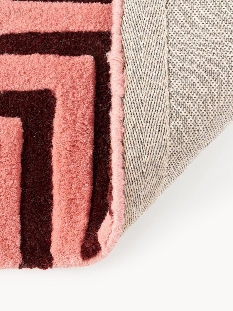 May include: Close-up of a pink and burgundy geometric patterned rug. The rug has a soft, textured surface with a light pink border and a neutral-colored backing. The design features a repeating pattern of burgundy lines.
