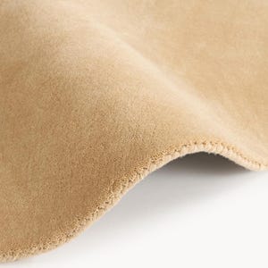 May include: Close-up of a section of tan fabric with a soft, velvety texture. The fabric is folded, revealing a glimpse of the underside and the stitched edge. The color is a warm, neutral tone, suitable for various textile applications.