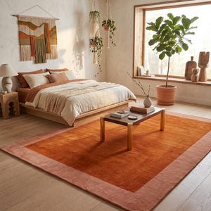 May include: A rectangular area rug featuring a deep burnt orange center panel framed by a lighter terracotta border. The rug is placed on a light-toned wooden floor in a room containing a bed, a low coffee table, and a large potted plant.