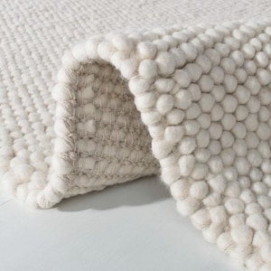 Puede incluir: Primer plano de una alfombra color crema con un diseño texturizado y en bucle. La alfombra está doblada, revelando el reverso tejido. La superficie tiene una textura suave y con nudos, creando un aspecto acogedor y acogedor.