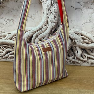 May include: A hobo-style shoulder bag featuring a vertical striped pattern in shades of yellow, red, purple, and light blue. The bag includes a wide shoulder strap, a top zip closure, and a small rectangular brown leather-like brand label.