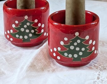 May include: Two cylindrical red ceramic candle holders, each featuring a green Christmas tree graphic with white polka dots. Each holder contains a single, tall, tapered, light brown candle with a visible white wick at the top.