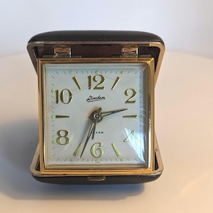 Puede incluir: Una taza de cerámica blanca con un asa negra y el texto "But First, Coffee" en negrita negra. La taza se encuentra sobre un fondo borroso, sugiriendo un ambiente acogedor.