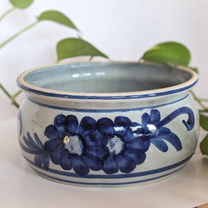 以下が含まれることがあります： 内側と外側が水色の丸い陶器の植木鉢。外側には、青い花のハンドペイントのデザインが施されています。背景には、緑の植物が写っています。