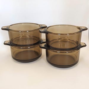 May include: Four vintage brown glass casserole dishes with handles are stacked on a white surface. The dishes are round with a slightly flared rim and two handles. The glass has a warm, amber hue, suggesting they are oven-safe.