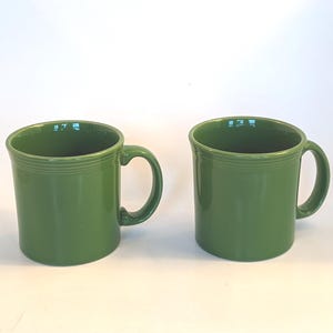 Puede incluir: Dos tazas de cerámica verdes con un diseño sencillo y clásico. Cada taza tiene un asa redondeada y un borde ligeramente ensanchado. Las tazas son de color verde sólido y vibrante y parecen estar hechas de material duradero.