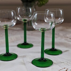 Op de afbeelding: Vier heldere wijnglazen met smaragdgroene stelen en voetjes staan op een wit tafelkleed. De glazen hebben een klassiek ontwerp, met een afgeronde kelk en een lange, slanke steel. De groene kleur voegt een vleugje elegantie toe.