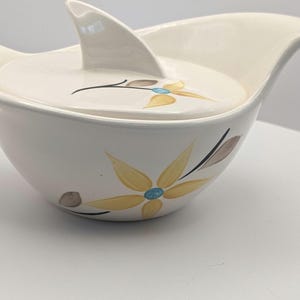 May include: Cream-coloured ceramic sugar bowl with a lid and a floral design. The bowl has a unique shape with a handle on each side and a fin-shaped handle on the lid. The floral design features yellow, brown, and blue accents.