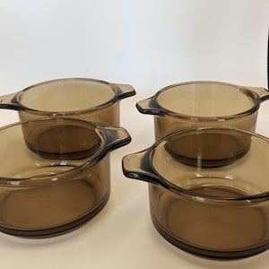 May include: Four amber-colored glass bowls with handles. The bowls are round with a slightly flared shape and are nested together. The glass has a warm, transparent hue, and the handles are a darker shade of brown.