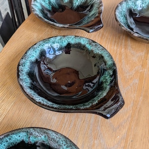 Può includere: Un set di tre ciotole rotonde in ceramica con interno marrone scuro lucido e bordo testurizzato con smalto turchese e nero maculato. Ogni ciotola presenta una piccola impugnatura integrata su un lato, creando una forma funzionale.