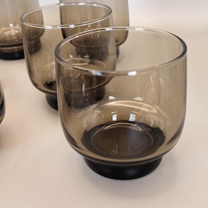May include: A collection of six smoked-glass tumblers. The glasses have a rounded bowl design and a dark, circular base. They are displayed on a light surface. Each glass is approximately 8 cm in height.