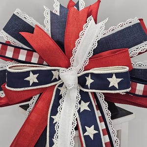 May include: A decorative bow featuring red, white, and blue ribbons. The bow has a patriotic theme with white stars on a navy blue ribbon and red and white striped ribbon accents. White lace trim edges the ribbons.