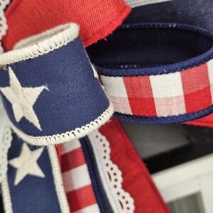 May include: Close-up of patriotic ribbon with red, white, and blue colors. The ribbon features a navy blue section with white embroidered stars, red and white stripes, and a red gingham pattern. White lace trim adds a delicate touch.