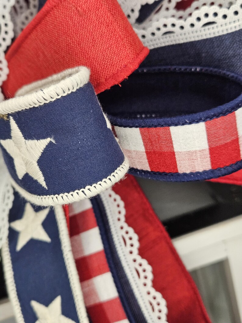 May include: Close-up of patriotic ribbon with red, white, and blue colors. The ribbon features a navy blue section with white embroidered stars, red and white stripes, and a red gingham pattern. White lace trim adds a delicate touch.