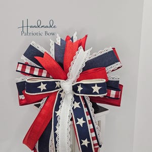 May include: A handmade patriotic bow featuring red, white, and blue ribbons. The bow includes ribbons with white stars on a navy background, red and white stripes, and white lace trim. The text "Handmade Patriotic Bow" is visible.