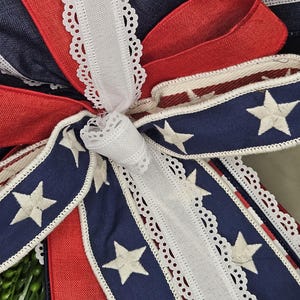 May include: Close-up of a decorative bow featuring patriotic colors. The bow has red, white, and blue ribbons. The blue ribbons are adorned with white stars. White lace trim edges the ribbons, creating a festive and detailed design.