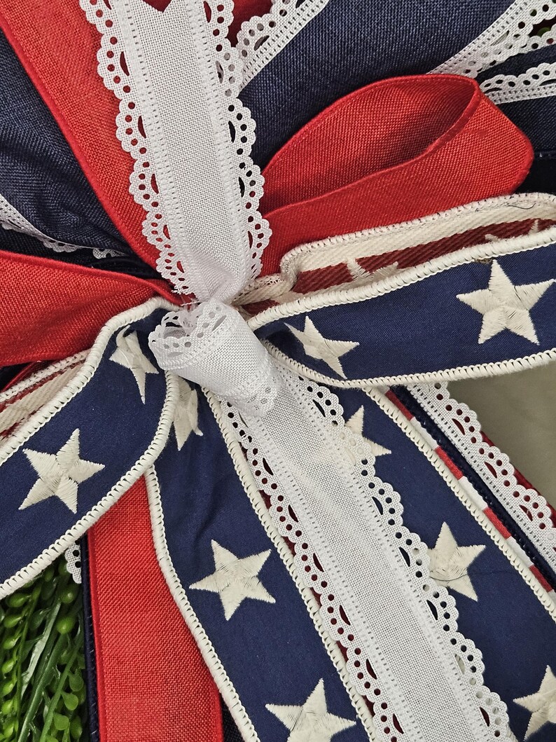 May include: Close-up of a decorative bow featuring patriotic colors. The bow has red, white, and blue ribbons. The blue ribbons are adorned with white stars. White lace trim edges the ribbons, creating a festive and detailed design.