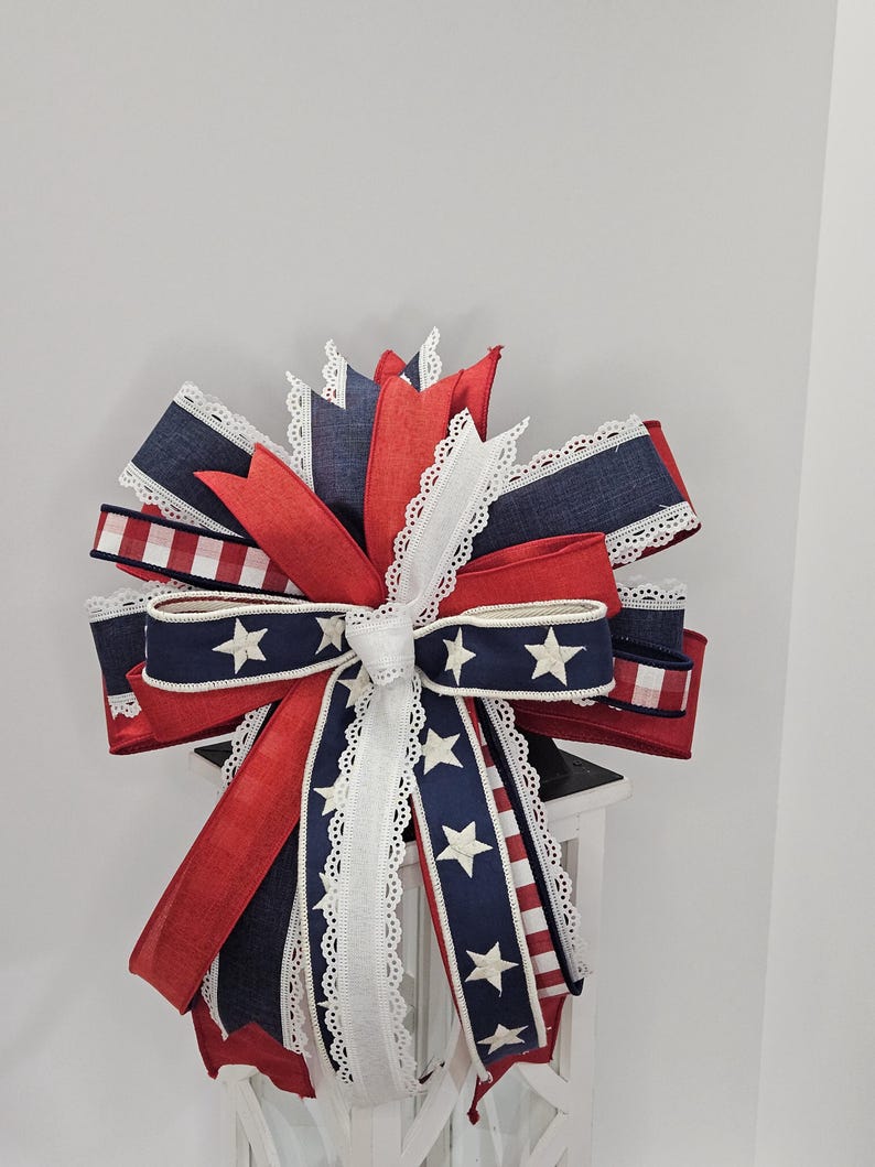 May include: A decorative bow featuring red, white, and blue ribbons. The bow has a patriotic theme with white stars on a navy blue ribbon and red and white striped ribbon accents. White lace trim edges the ribbons.