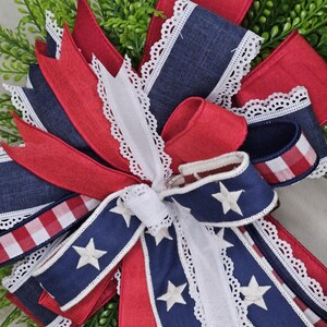 May include: A patriotic bow featuring red, white, and blue ribbons. The bow includes navy blue ribbons with white stars, red ribbons, and white lace trim. The bow is set against a backdrop of green foliage.