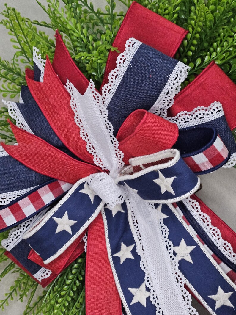 May include: A patriotic bow featuring red, white, and blue ribbons. The bow includes navy blue ribbons with white stars, red ribbons, and white lace trim. The bow is set against a backdrop of green foliage.