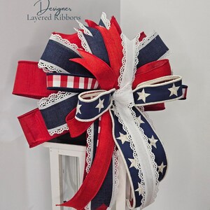 May include: A decorative bow featuring red, white, and navy blue ribbons. The bow includes ribbons with white stars, lace trim, and a gingham pattern. The text "Designer Layered Ribbons" is visible in the upper left corner.