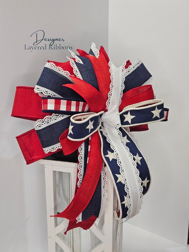 May include: A decorative bow featuring red, white, and navy blue ribbons. The bow includes ribbons with white stars, lace trim, and a gingham pattern. The text "Designer Layered Ribbons" is visible in the upper left corner.