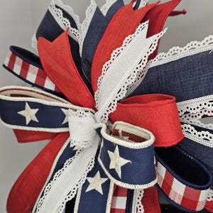 May include: A patriotic bow featuring red, white, and blue ribbons. The bow includes navy blue ribbons with white stars, red ribbons, and white lace-edged ribbons. A red and white checkered ribbon is also present.