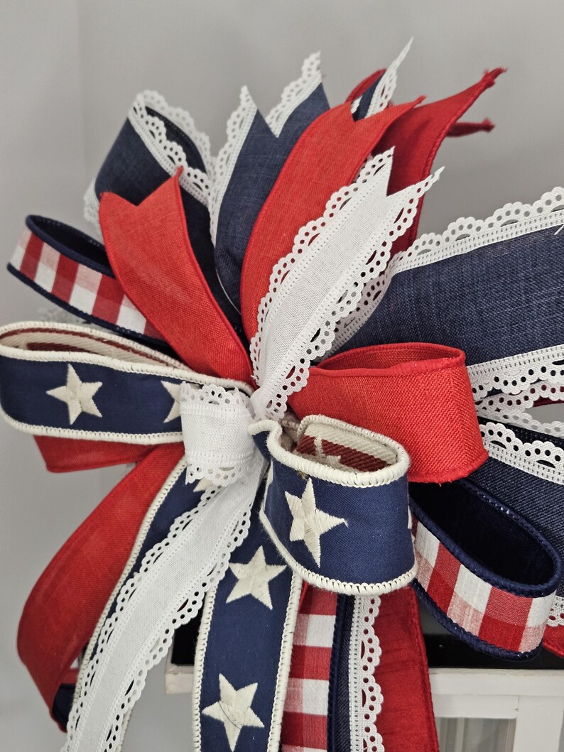 May include: A patriotic bow featuring red, white, and blue ribbons. The bow includes navy blue ribbons with white stars, red ribbons, and white lace-edged ribbons. A red and white checkered ribbon is also present.