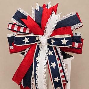 May include: A decorative bow featuring red, white, and blue ribbons, accented with white lace trim and star patterns. The bow is designed with patriotic colors and is likely used for festive decorations.