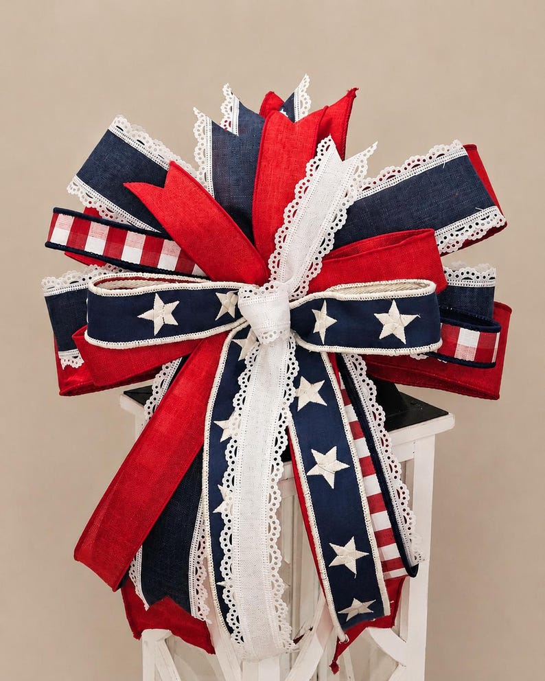May include: A decorative bow featuring red, white, and blue ribbons, accented with white lace trim and star patterns. The bow is designed with patriotic colors and is likely used for festive decorations.
