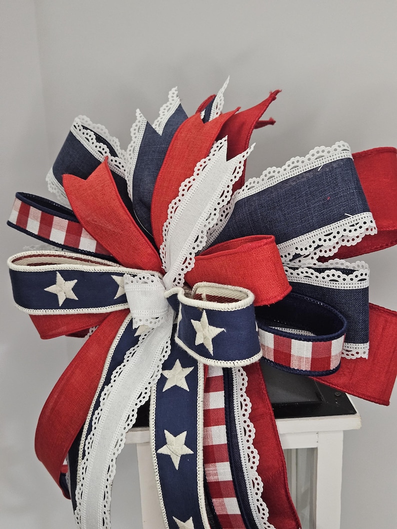 May include: A patriotic bow featuring red, white, and blue ribbons. The bow includes ribbons with white stars on a navy background, red and white gingham, and white lace trim. This decorative bow is perfect for Independence Day.