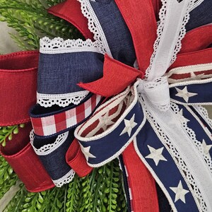 May include: A patriotic wreath with red, white, and blue ribbons. The wreath features denim, star-spangled, and gingham ribbons, accented with white lace trim. Greenery forms the base, creating a festive, decorative piece.