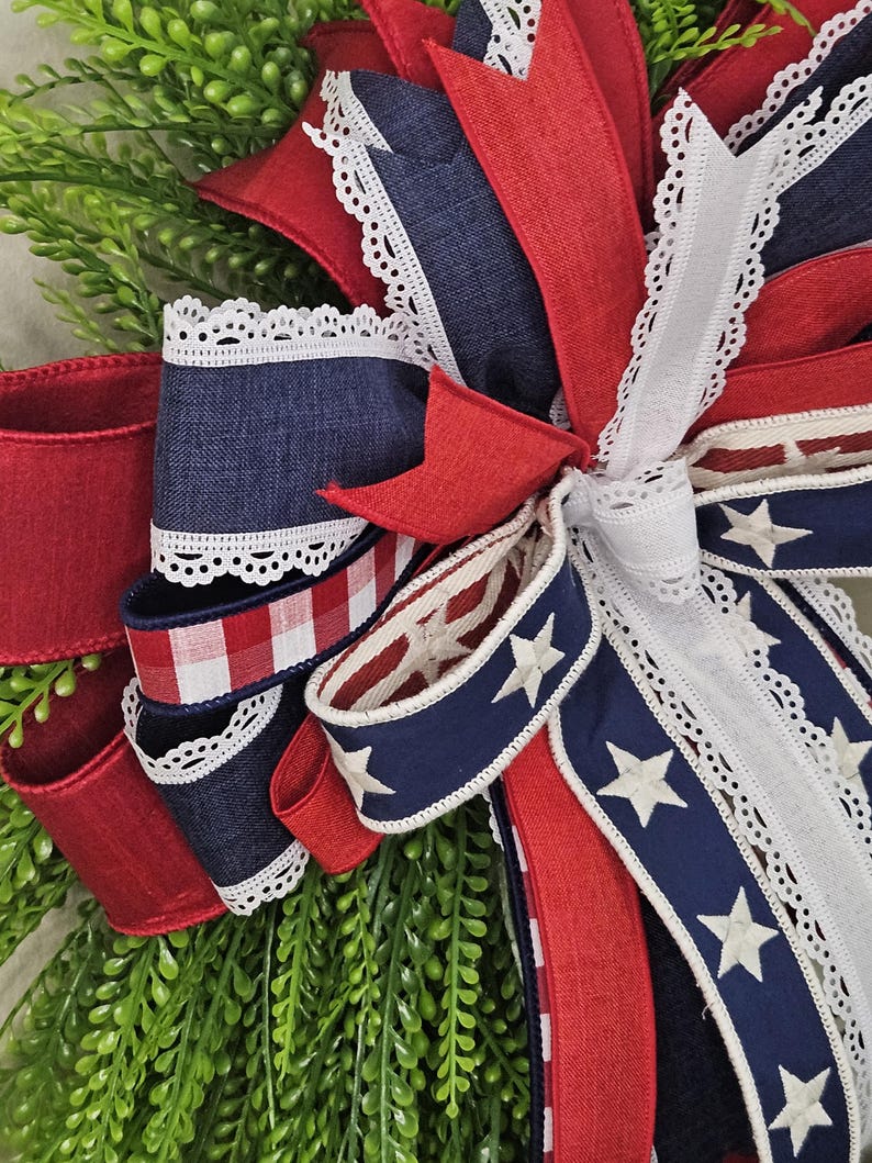 May include: A patriotic wreath with red, white, and blue ribbons. The wreath features denim, star-spangled, and gingham ribbons, accented with white lace trim. Greenery forms the base, creating a festive, decorative piece.