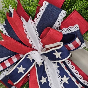 May include: A patriotic bow featuring red, white, and blue ribbons. The bow includes denim-like blue ribbons with white stars, red ribbons, and white ribbons with a delicate lace trim. The bow is set against a backdrop of green foliage.