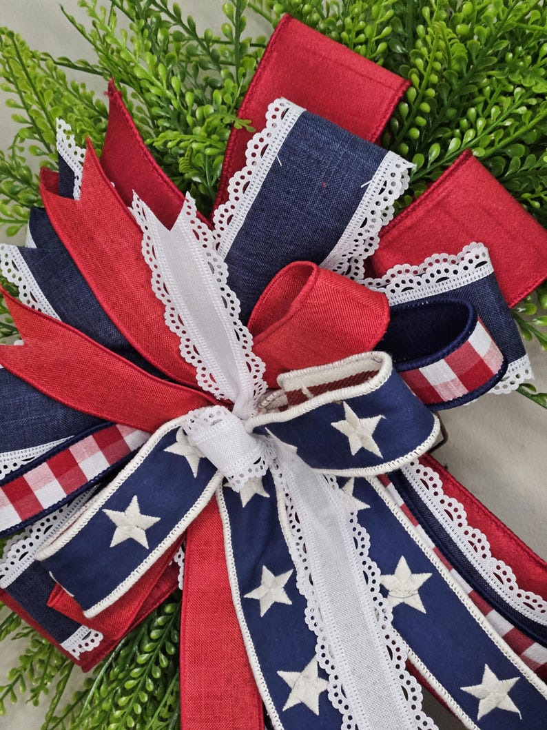 May include: A patriotic bow featuring red, white, and blue ribbons. The bow includes denim-like blue ribbons with white stars, red ribbons, and white ribbons with a delicate lace trim. The bow is set against a backdrop of green foliage.
