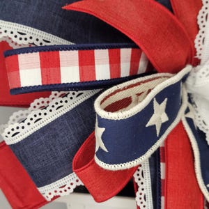 May include: Close-up of a decorative bow featuring patriotic colors. The bow incorporates red, white, and blue ribbons with various patterns, including gingham, stars, and lace trim. The design evokes a festive, celebratory theme.
