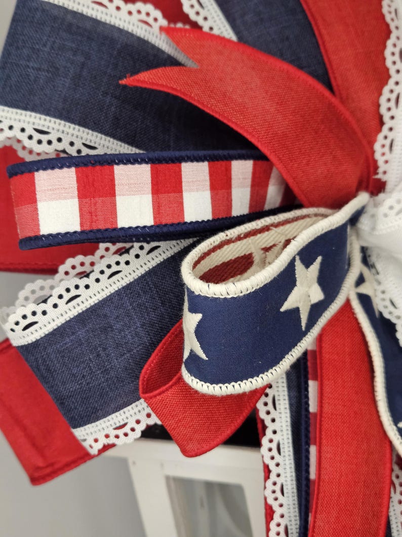 May include: Close-up of a decorative bow featuring patriotic colors. The bow incorporates red, white, and blue ribbons with various patterns, including gingham, stars, and lace trim. The design evokes a festive, celebratory theme.