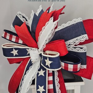 May include: A patriotic bow featuring red, white, and blue ribbons with white lace trim and star accents. The bow is designed with a mix of solid and patterned fabrics, perfect for Memorial Day and 4th of July decorations.