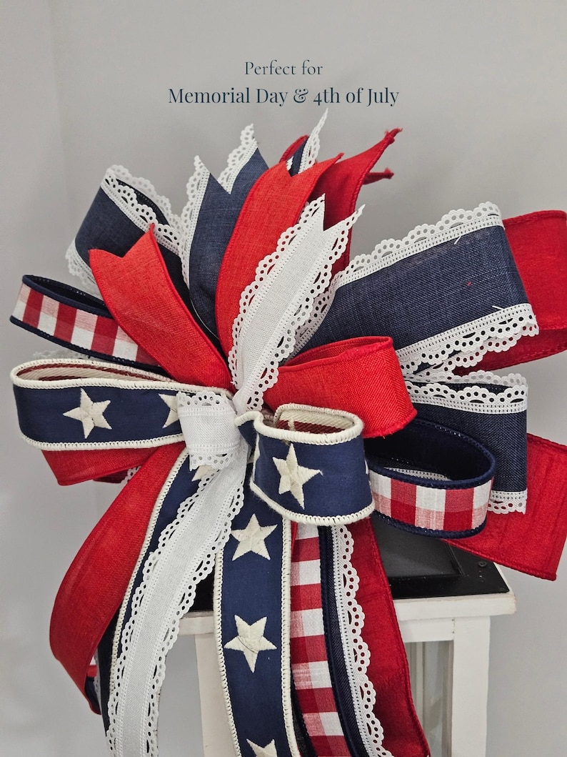 May include: A patriotic bow featuring red, white, and blue ribbons with white lace trim and star accents. The bow is designed with a mix of solid and patterned fabrics, perfect for Memorial Day and 4th of July decorations.