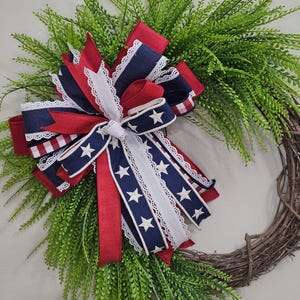 May include: A decorative wreath featuring a base of interwoven brown twigs and lush green foliage. A large bow made of red, white, and blue ribbons with star patterns and lace trim is the focal point, creating a patriotic theme.