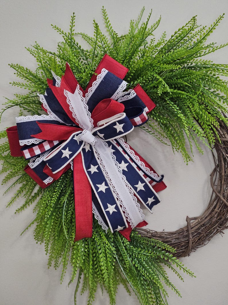 May include: A decorative wreath featuring a base of interwoven brown twigs and lush green foliage. A large bow made of red, white, and blue ribbons with star patterns and lace trim is the focal point, creating a patriotic theme.