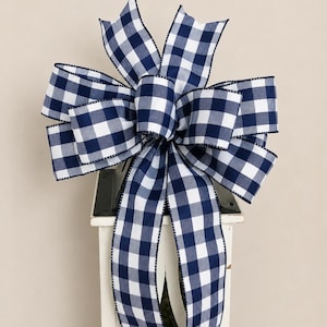 May include: A decorative bow crafted from fabric featuring a navy blue and white buffalo check pattern. The bow has multiple loops and two long, trailing tails that hang downward, with edges finished in a dark, thin stitched border detail.