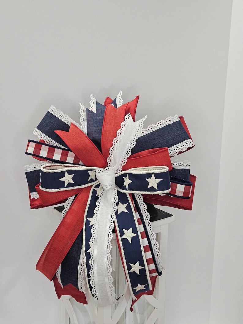 May include: A decorative bow featuring red, white, and blue ribbons. The bow has a patriotic theme with star and stripe patterns. White lace trim edges the ribbons, adding a delicate touch. The bow is likely for festive decor.