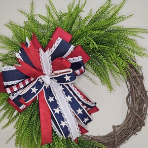 May include: A patriotic wreath featuring a grapevine base, adorned with lush green foliage and a large bow. The bow incorporates red, white, and blue ribbons, some with star patterns and lace trim, creating a festive and decorative piece.